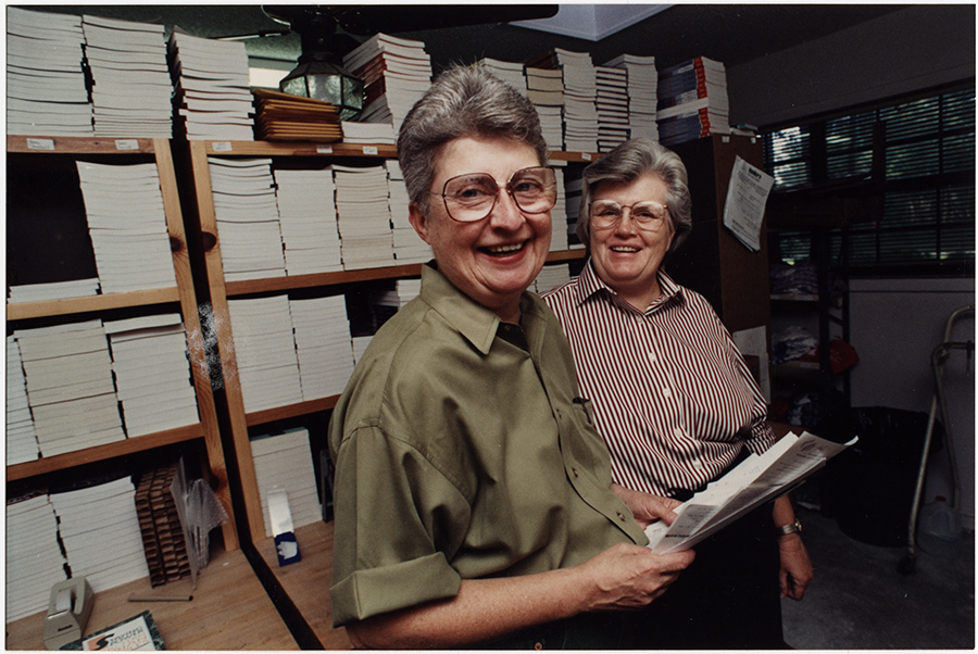 Two women with short grey hair and glasses smile at the camera. There are shelves full of books behind them. 