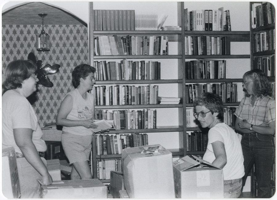 A black and white vintage photo featuring four women hard at work packing books. Shelves full of books are behind them.