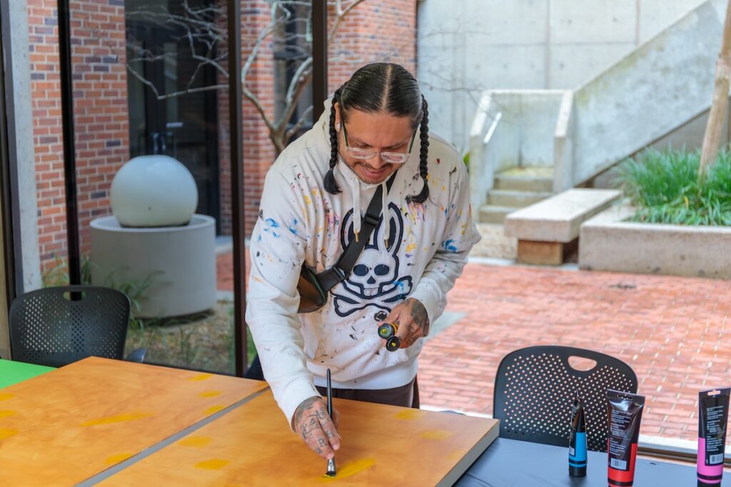 A man in a white sweatshirt with black braised hair leans over to paint on a canvas. 
