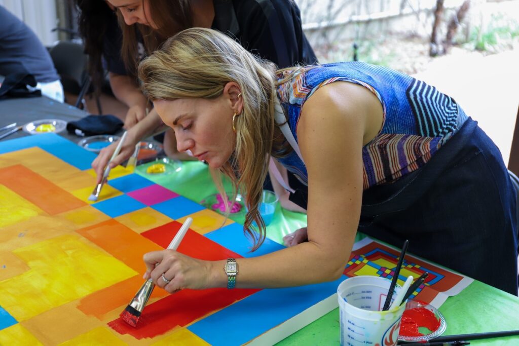 A woman leans over to paint on a canvis with a bright design. 